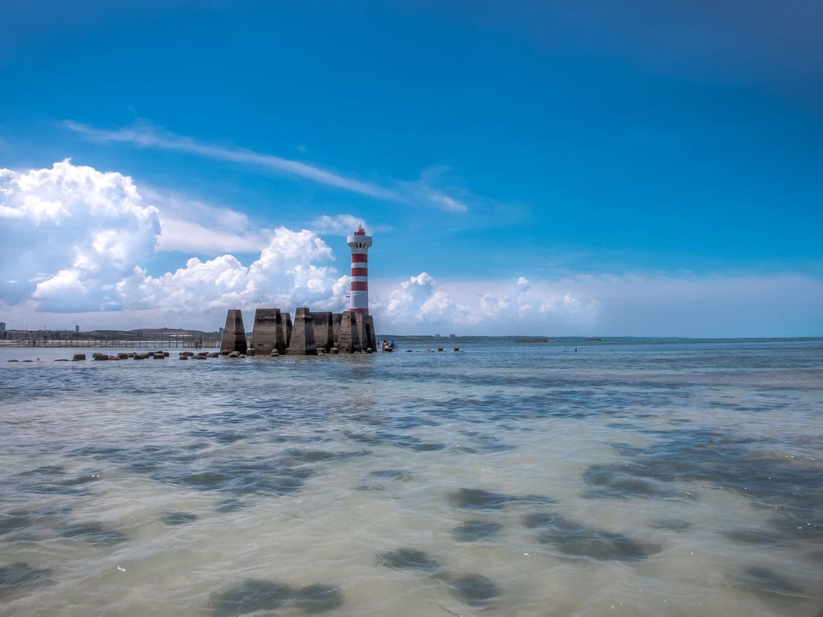 Águas rasas e recifes parcialmente expostos diante do Farol da Ponta Verde, em Maceió. Ilustrando artigo Piscinas naturais de Maceió: por que dependem da maré?