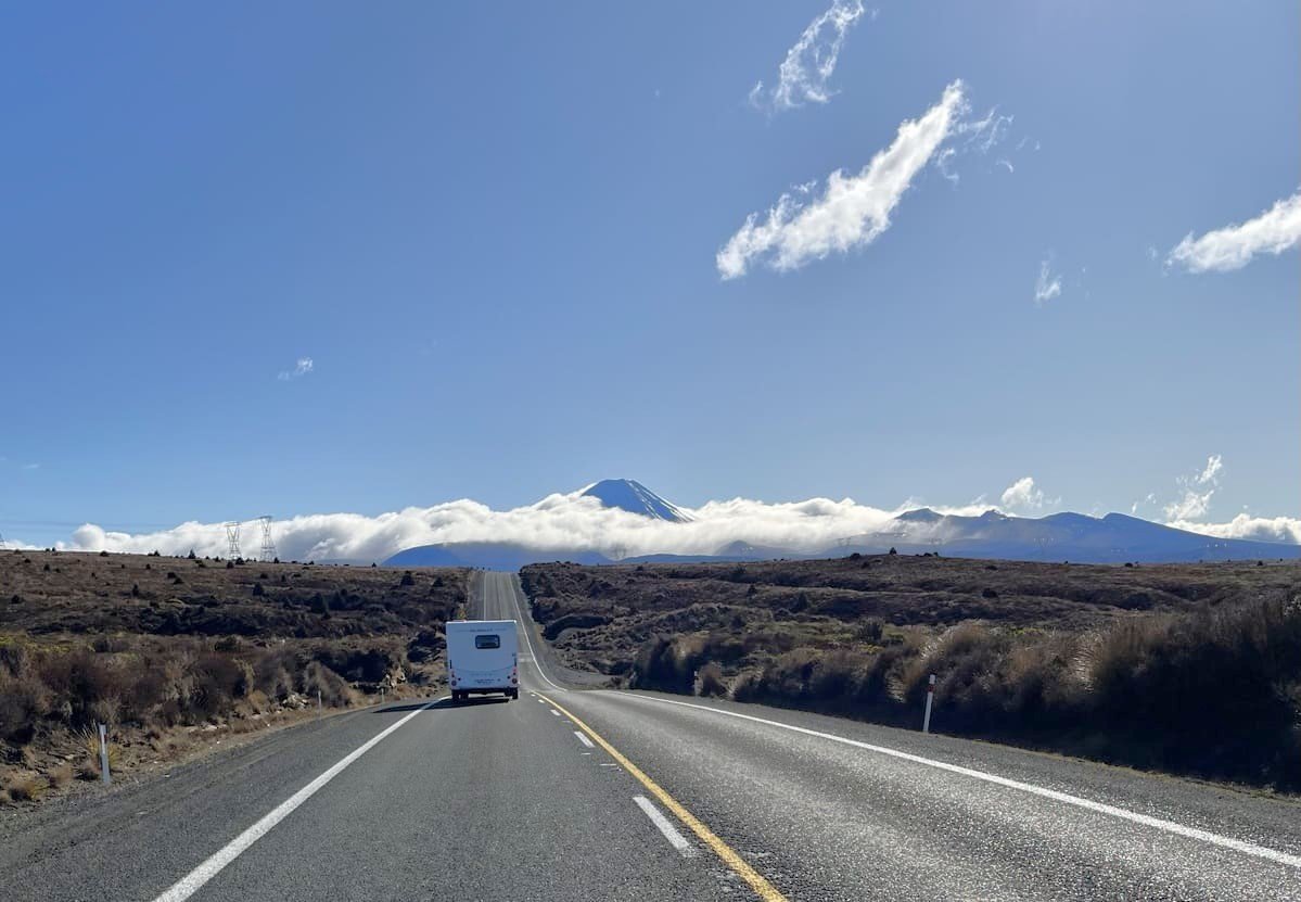 Estrada com veículo e Ngauruhoe parcialmente encoberto por nuvens ao fundo. Ilustrando artigo Ilha Norte da Nova Zelândia: por que há tantos vulcões?