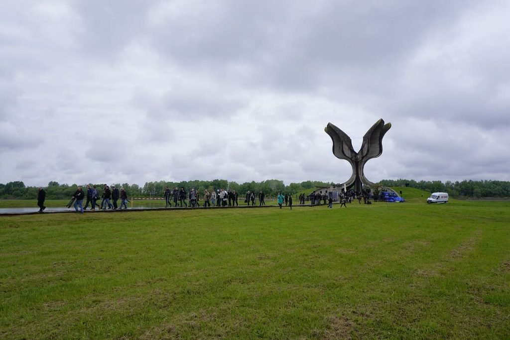 Pessoas no caminho de acesso ao Monumento da Flor de Jasenovac, entre gramados e uma superfície de água, sob céu nublado. Ilustrando artigo Monumentos de concreto na Europa: por que estão ali?