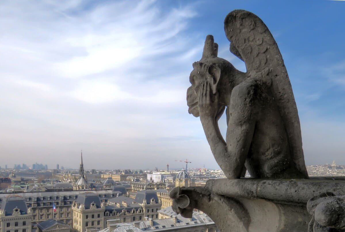 Stryge de pedra nas torres de Notre-Dame, com edifícios de Paris ao fundo. Ilustrando artigo Histórias de Paris: o que há de real nos cenários?
