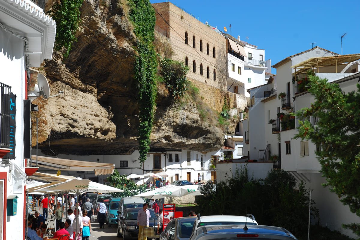 Fachadas brancas sob uma saliência rochosa em Setenil, com pessoas, mesas e veículos na rua. Ilustrando artigo Construções na rocha na Espanha: como o terreno é usado?