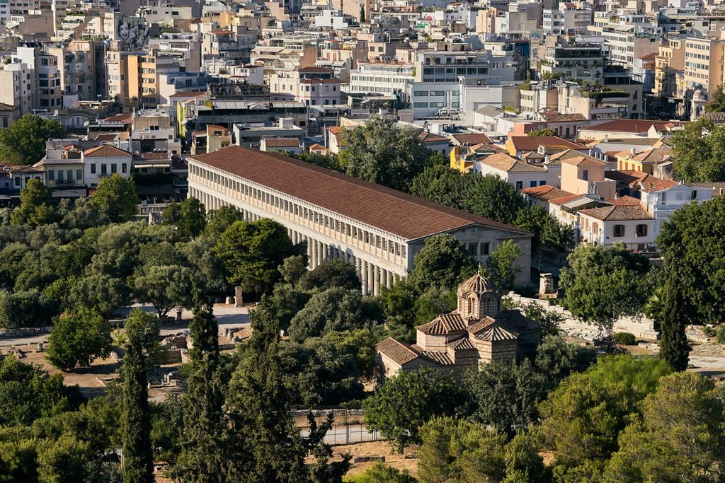 Estoa de Átalo entre árvores e vestígios da Ágora de Atenas, com edifícios urbanos ao fundo.