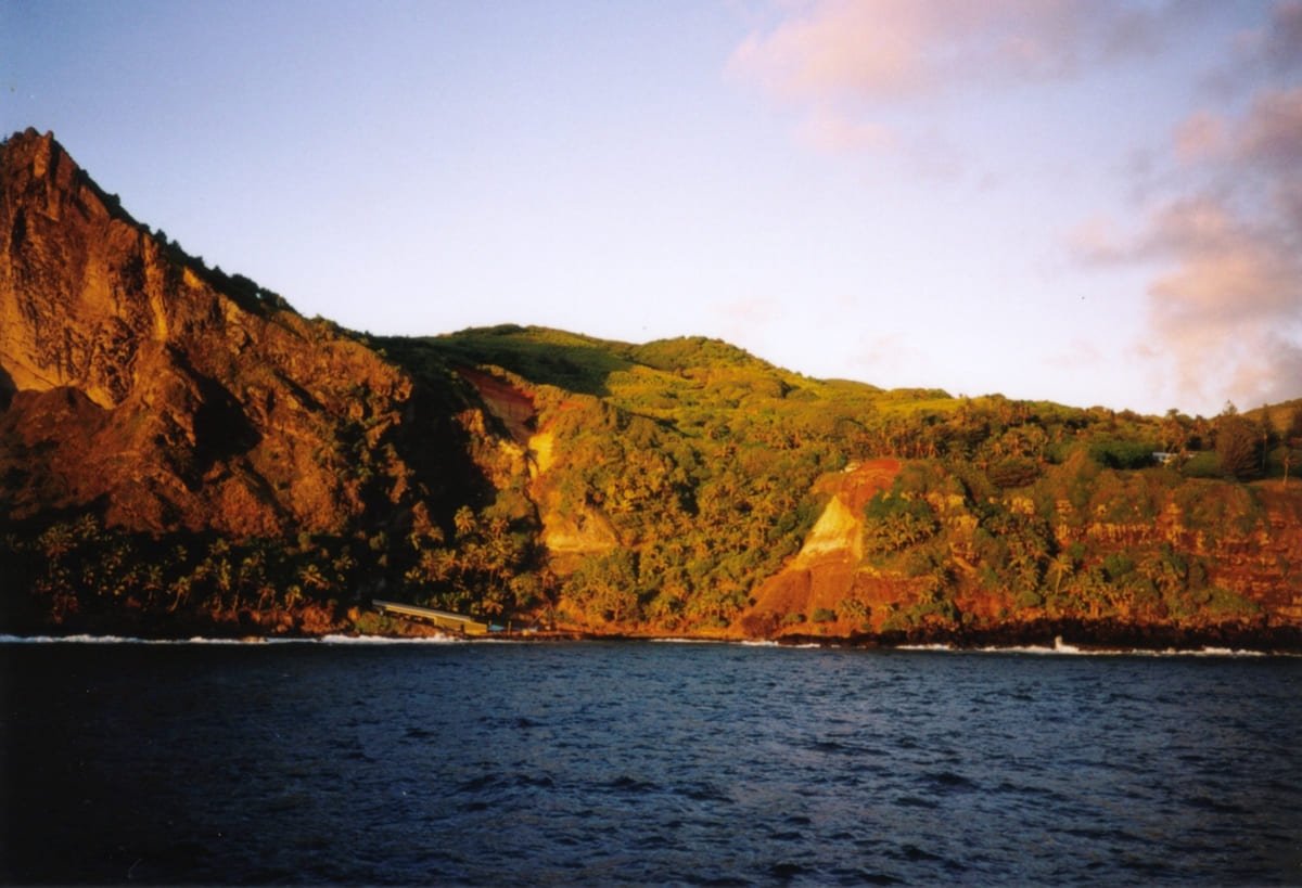 Bounty Bay, em Pitcairn, com costa rochosa, vegetação e estruturas junto ao mar. Ilustrando artigo Ilhas remotas da Oceania: por que é tão difícil chegar?
