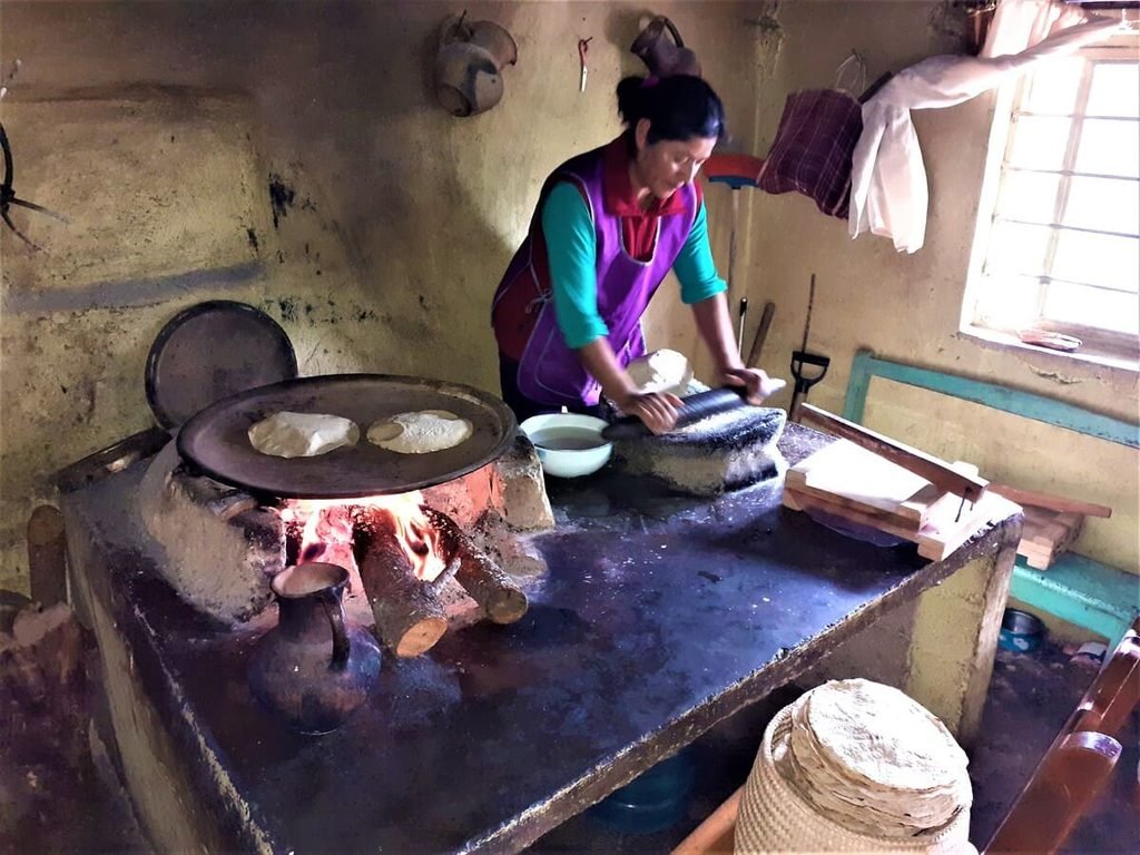 Mulher preparando tortillas, com massa sobre um metate e comal de barro sobre o fogão.