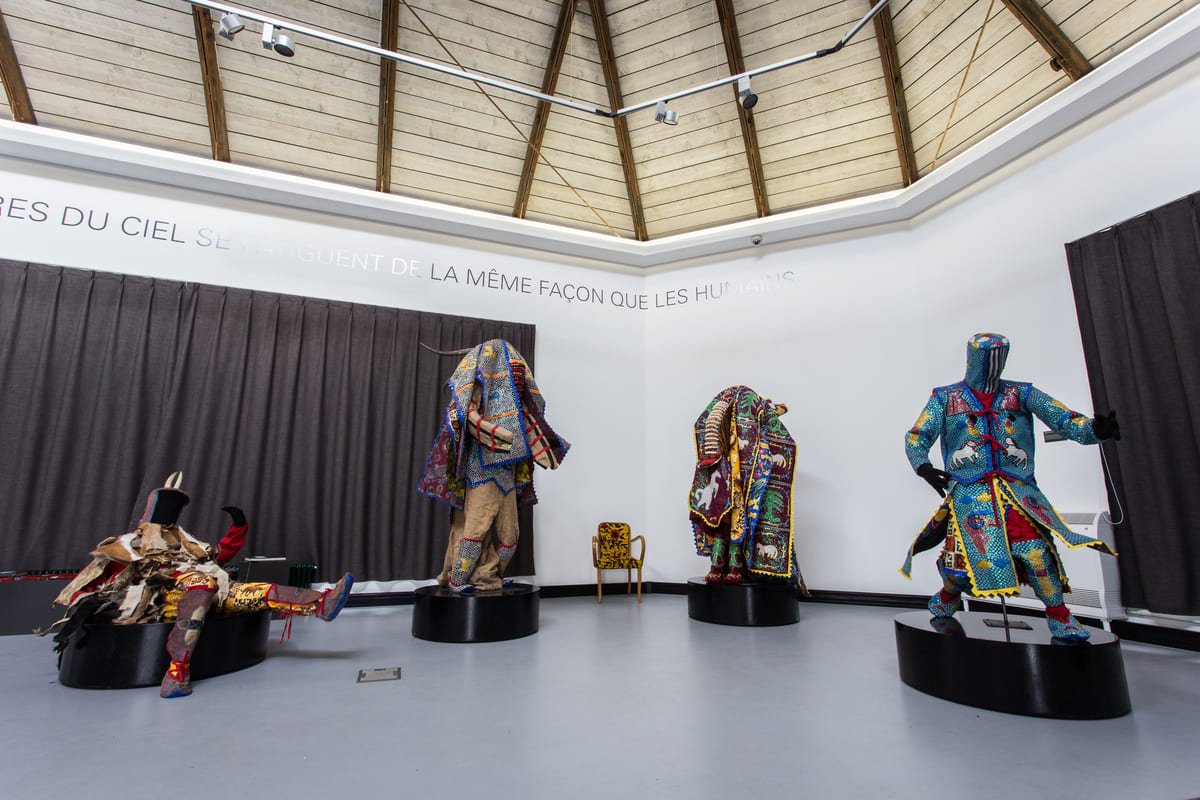 Quatro conjuntos de trajes Egungun com tecidos coloridos e ornamentos em exposição no Château Vodou. Ilustrativo ao artigo Máscaras africanas: o que a peça sozinha não revela?