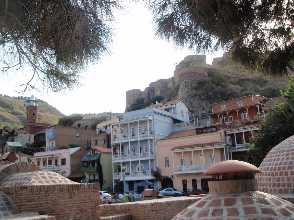 Cúpulas dos banhos de Abanotubani entre edifícios, com a encosta de Narikala ao fundo, ilustrando artigo Banhos de Tbilisi: por que se concentram em Abanotubani?