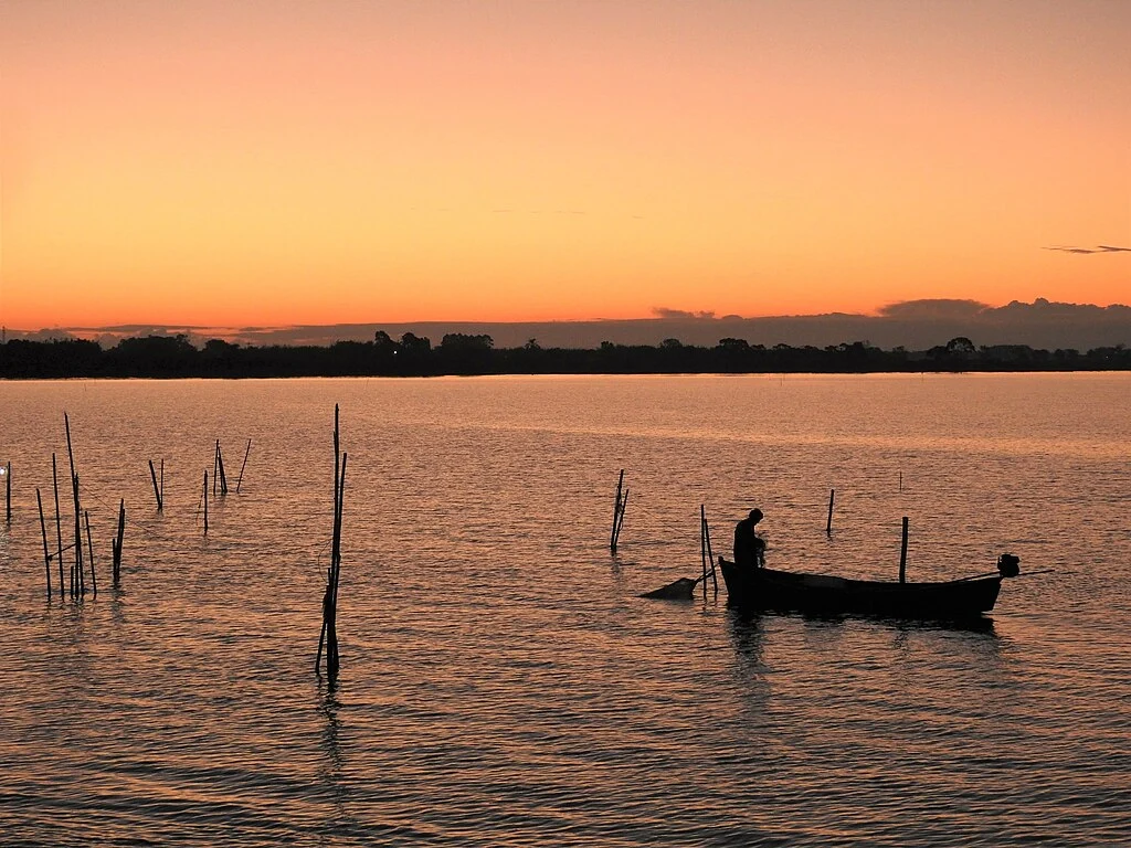 Foto em contraluz, no por do sol com pescador sobre embarcação parada sobre águas da Lagoa dos Patos, um dos 10 Lagos Gigantes do Brasil.