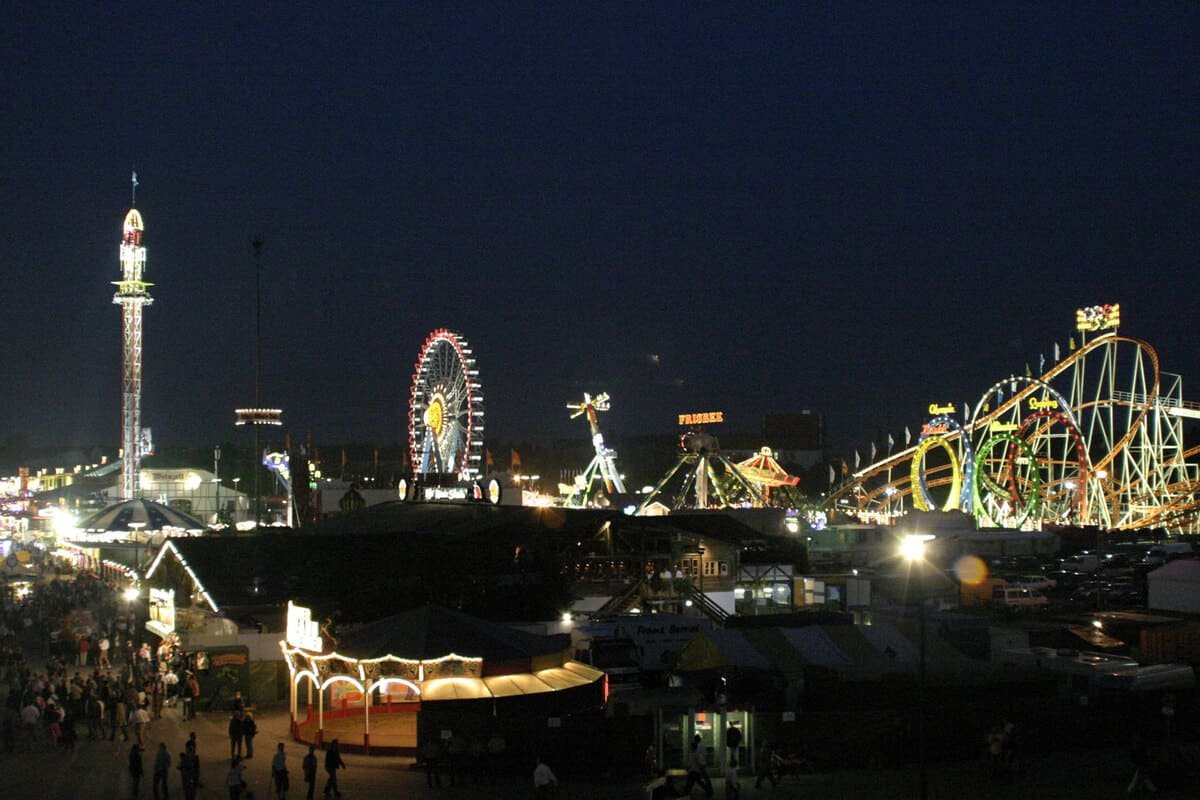 Vista noturna da Oktoberfest de Munique, em setembro de 2005, com barracas e atrações iluminadas na Theresienwiese. Fotografia: Bjs, via Wikimedia Commons, disponibilizada sob CC0 1.0.